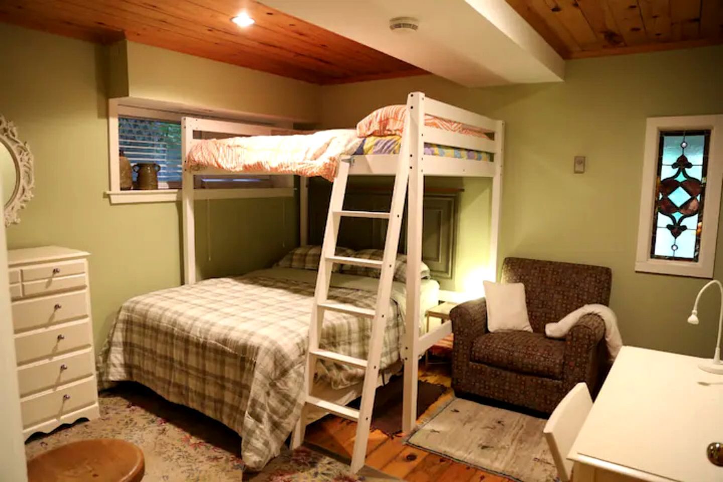 A bunk bed with a ladder, a chair, a desk, and a dresser in a room with wooden ceiling beams and a window with blinds.