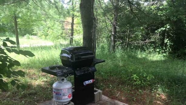 A portable camping stove with a gas canister and a black pot on a concrete base in a wooded area. - Tiny House Rental in Markdale Perfect for a Couple's Retreat in Ontario (Markdale)
