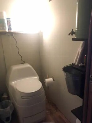 Un baño pequeño con un inodoro, un balde y un estante con objetos.