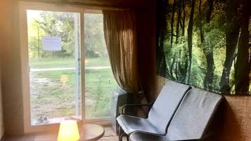 A room with a sofa, a lamp, a window with a curtain, and a view of a forest.