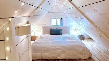 Ein Schlafzimmer im Lofts-Stil mit schrägem Dach, einem Bett mit gestreifter Bettdecke, einem Fenster mit Vorhängen und einem Nachttisch mit Lampe.