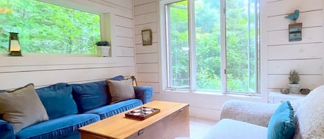 Ein Zimmer mit einem blauen Sofa, einem weißen Sessel, einer hölzernen Kommode und großen Fenstern mit Blick auf Grünflächen.