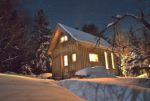 A cabin with a snow-covered roof, wooden walls, and a window with a visible nameplate.