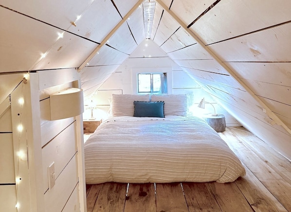 Un dormitorio tipo loft con techo inclinado, cama con colcha a rayas, ventana con cortinas y mesita de noche con lámpara.
