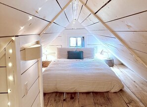 A loft bedroom with a sloped ceiling, a bed with a striped bedspread, a window with curtains, and a nightstand with a lamp. - Charming Cabin Rental on Deer Lake in Ontario, Canada (South River)