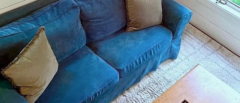 Un sofá azul con cojines, una mesa de centro de madera con un juego, y vistas al exterior entre la vegetación.