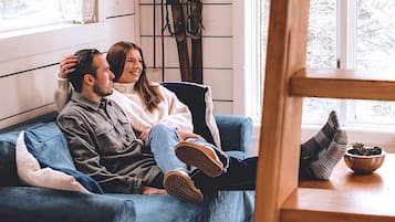 Una pareja sentada en un sofá azul en una habitación con una ventana, una mesa de madera con un cuenco y esquíes apoyados en la pared.