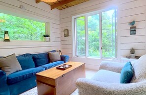 A room with a blue sofa, a white armchair, a wooden chest of drawers, and large windows with a view of greenery. - Charming Cabin Rental on Deer Lake in Ontario, Canada (South River)