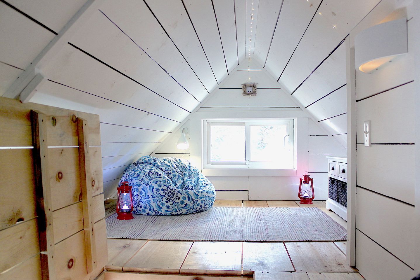 A room with a sloped ceiling, a window, a bed with patterned bedding, and two lanterns.