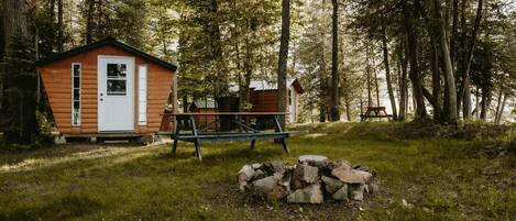 A rustic cabin with a wooden exterior, a small porch, and a picnic table in a forested area.