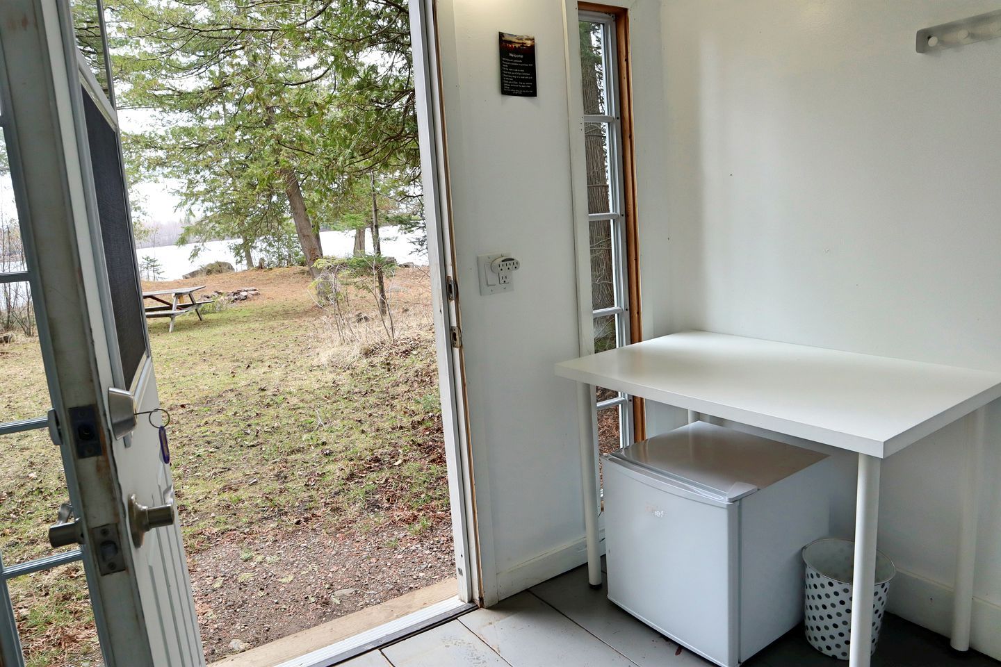 A room with a white desk, a small refrigerator, and a door leading to an outdoor area with trees and a picnic table.
