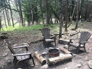 Entorno de bosque con tres sillas Adirondack dispuestas alrededor de una fogata.