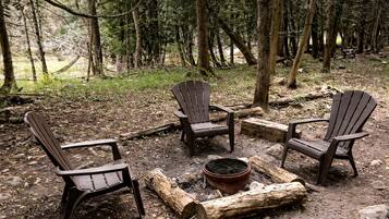 A campsite with three Adirondack chairs arranged around a campfire, surrounded by trees and natural terrain.