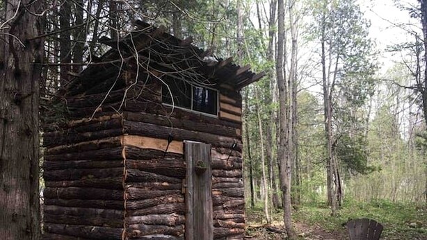 Una cabaña rústica de troncos con una terraza de madera y una sola puerta, rodeada de bosque.