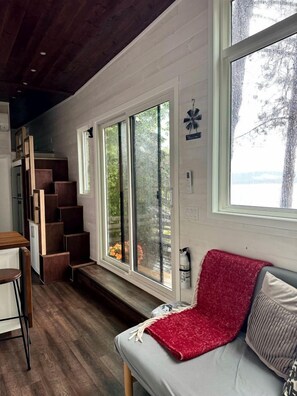 A room with a wooden staircase, a bench with a red blanket, and large windows with a view of trees. - Gorgeous Tiny House Nestled Amidst Trees and Fire Pit Provided in Petawawa, Ontario (Petawawa)