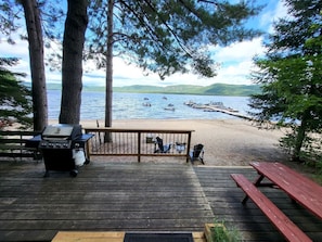 A wooden deck with a grill, chairs, and a picnic table overlooking a lake with boats and hills in the background.