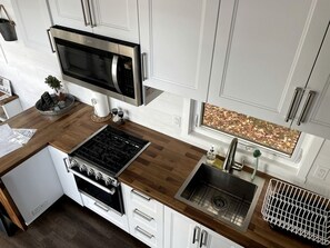 A modern kitchen with a microwave, stove, sink, and a window with a leaf pattern. - Gorgeous Tiny House Nestled Amidst Trees and Fire Pit Provided in Petawawa, Ontario (Petawawa)