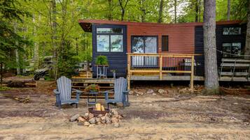 A cabin with a deck, two Adirondack chairs, a fire pit, and a forested backdrop.