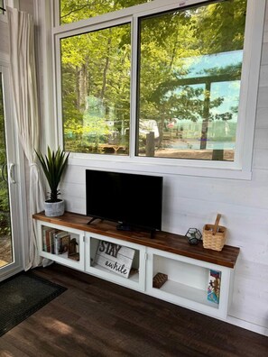 A room with a TV on a wooden shelf, a potted plant, a sign that says 'STAY awhile,' and a view of trees and a park through a large window.