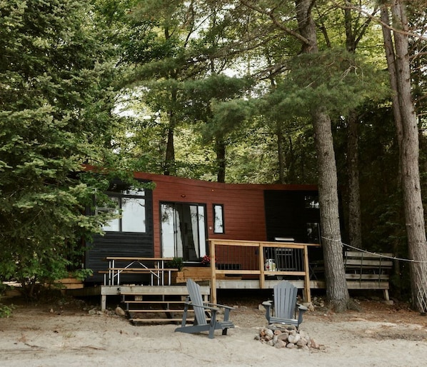 A cabin with a deck, two Adirondack chairs, and a wooden staircase leading to the entrance. - Gorgeous Tiny House Nestled Amidst Trees and Fire Pit Provided in Petawawa, Ontario (Petawawa)