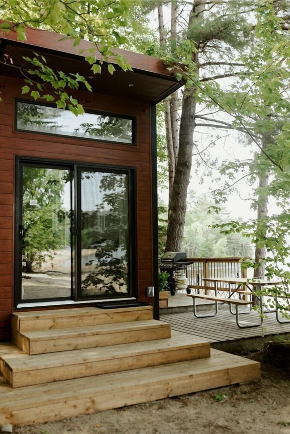 Une cabane en bois avec une porte en verre, une terrasse dotée d’un barbecue et des marches menant à l’entrée.