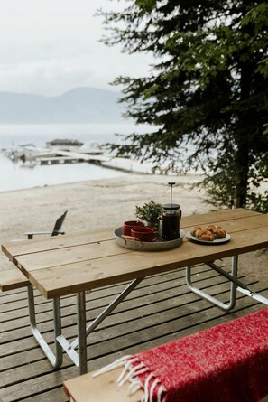A wooden picnic table with a coffee pot, red cups, and pastries on a deck overlooking a body of water.