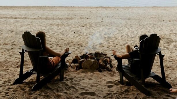 Dos sillas mirando a una fogata en una playa arenosa, con un lago y montañas al fondo.