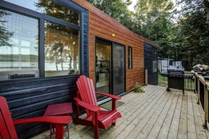 A modern wooden cabin with a deck, red Adirondack chairs, and a barbecue grill.