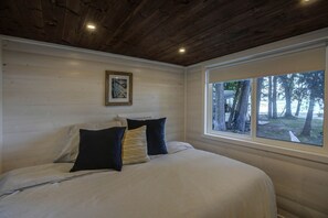 A bedroom with a wooden ceiling, a bed with white bedding, two pillows, a framed picture on the wall, and a window with a view of trees.