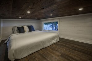 A bedroom with a large bed, wooden flooring, a window with a view of trees, and a white wall.