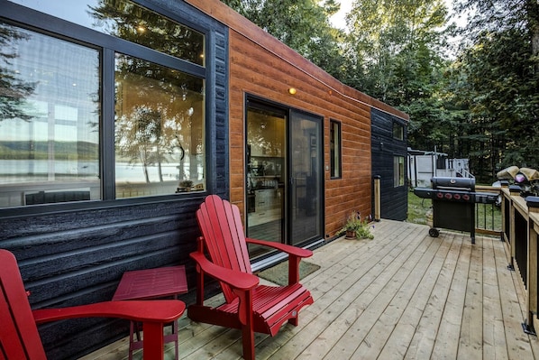 A modern wooden cabin with a deck, red Adirondack chairs, and a barbecue grill.