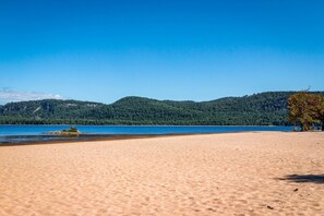 Ein Strand mit blauem Himmel, sandigem Ufer und einer bewaldeten Hügelkette im Hintergrund.