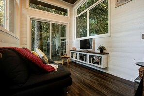 A living room with a black sofa, a TV on a white console, and large windows with a view of trees.