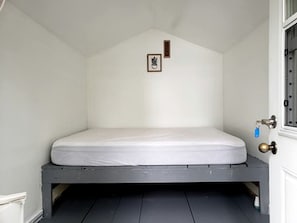 A small, narrow room with a single bed, a door with a handle, and a small framed picture on the wall.