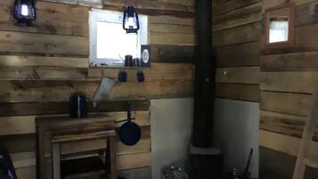 A rustic cabin interior with wooden walls, a window, a stove, a table with a kettle, and hanging lanterns.