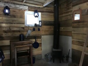 L’intérieur d’un chalet rustique, avec des murs en bois, une fenêtre, un poêle, une table avec une bouilloire et des lanternes suspendues.