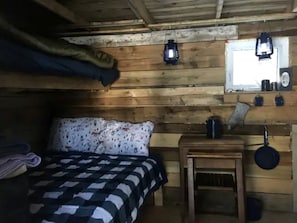 A wooden cabin room with a bed, a small table, a hanging lantern, and a window.
