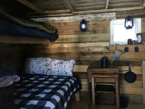 A wooden cabin room with a bed, a small table, a hanging lantern, and a window.