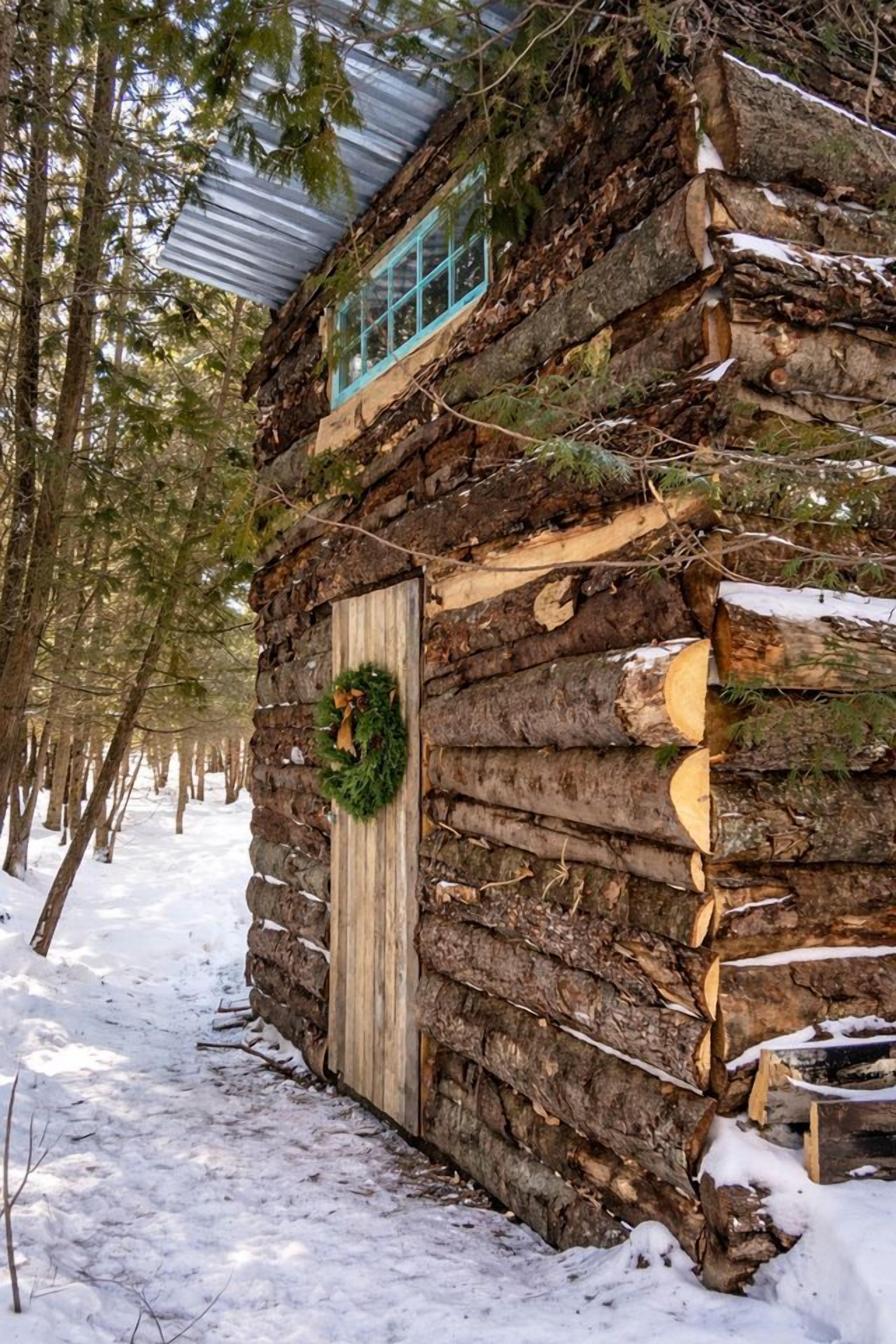 A log cabin with a wooden door and a wreath, surrounded by snow and trees.