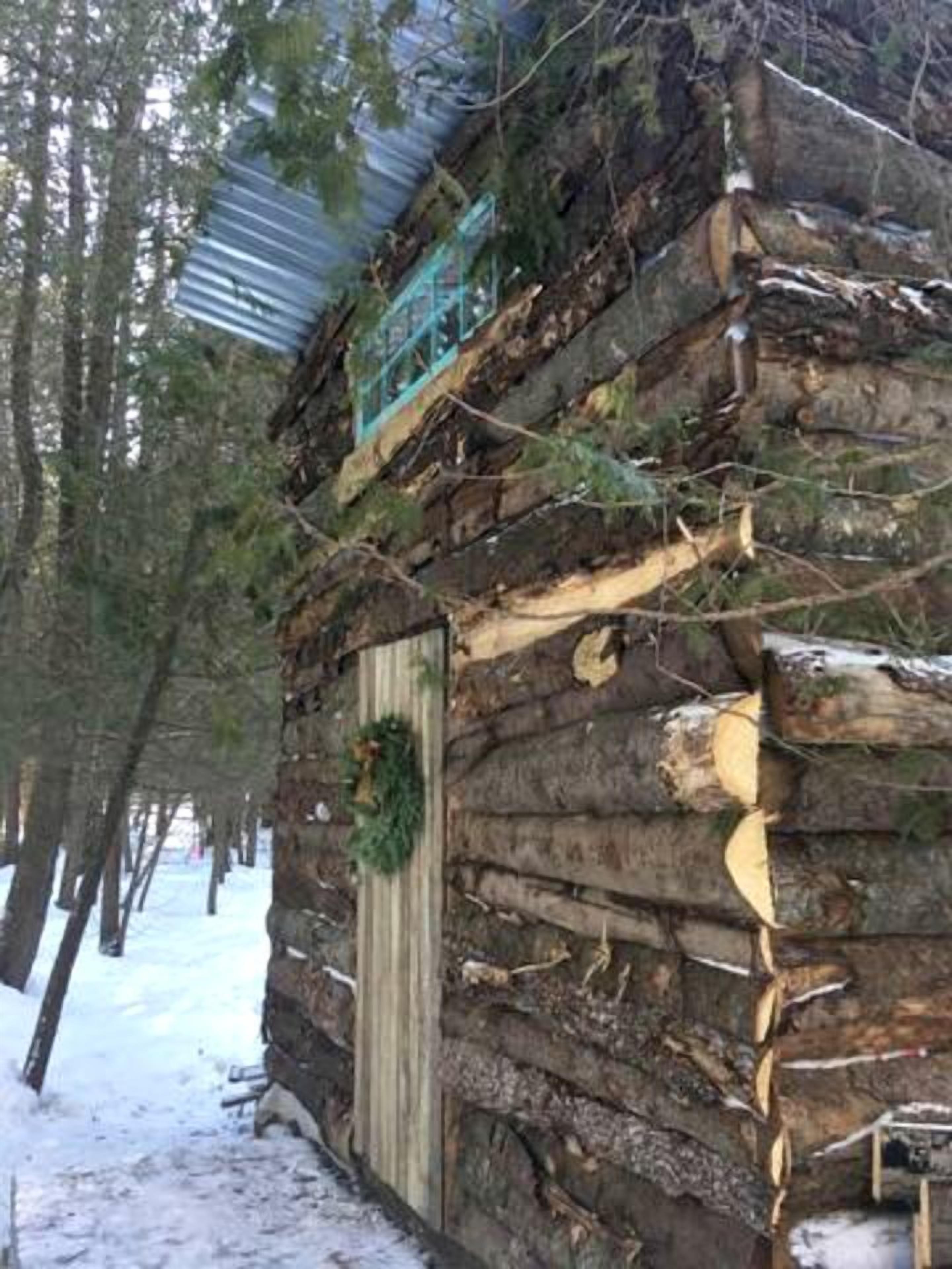 A rustic cabin with a wooden door and a wreath, surrounded by snow and trees.