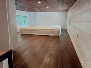 A room with wooden flooring, a mattress, a window, and a white wall.