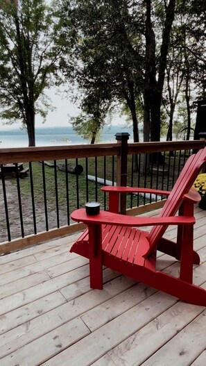 A red Adirondack chair on a wooden deck with a metal railing, overlooking a body of water and trees.