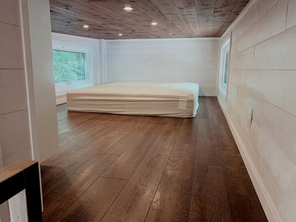 Una habitación con suelo de madera, un colchón, una ventana y una pared blanca.