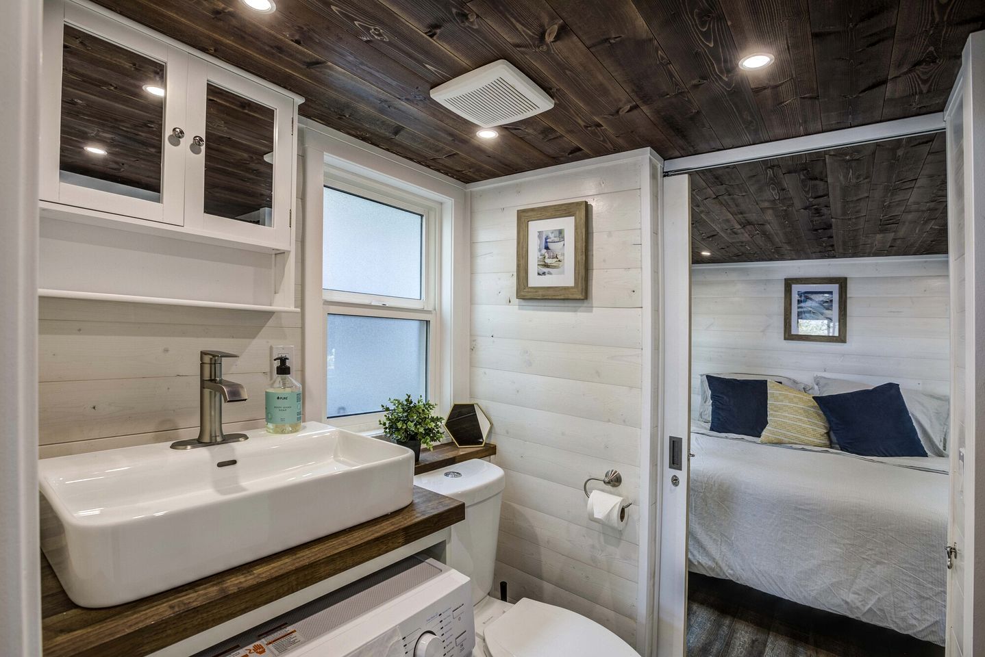 Une salle de bain compacte avec un lavabo blanc, des toilettes et un espace douche. Une fenêtre laisse entrer la lumière naturelle, et on aperçoit un lit avec des oreillers à travers une porte ouverte.