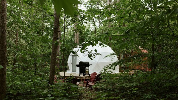 Ein kuppelförmiges Zelt im Wald mit Grill und Stühlen davor.