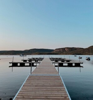 Eine hölzerne Steg führt ins ruhige Wasser, mit anlegenden Booten und einer hügeligen Landschaft im Hintergrund.
