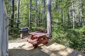A wooden deck with a picnic table and grill, surrounded by a forest.