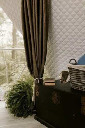 Une pièce avec un mur recouvert d’un tissu matelassé, une commode de couleur sombre, un panier en osier et un rideau.