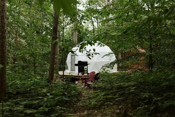 A dome-shaped tent in a forest with a grill and chairs outside.