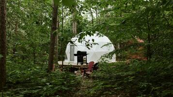 Une tente en forme de dôme, installée dans une forêt, avec un barbecue et des chaises à l’extérieur.